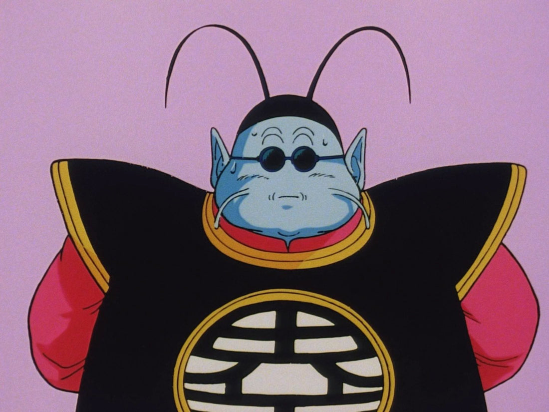 Portrait of King Kai from the front against a pink background: blue skin, pointed ears, round glasses, black antennae and a black tunic with a medallion.
